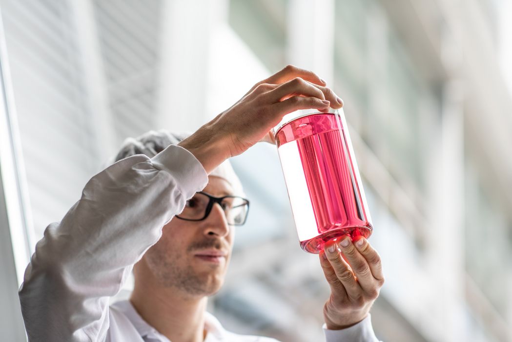 Wissenschaftler mit Schutzkleidung hält ein durchsichtiges Gefäß mit roter Flüssigkeit hoch und untersucht es.