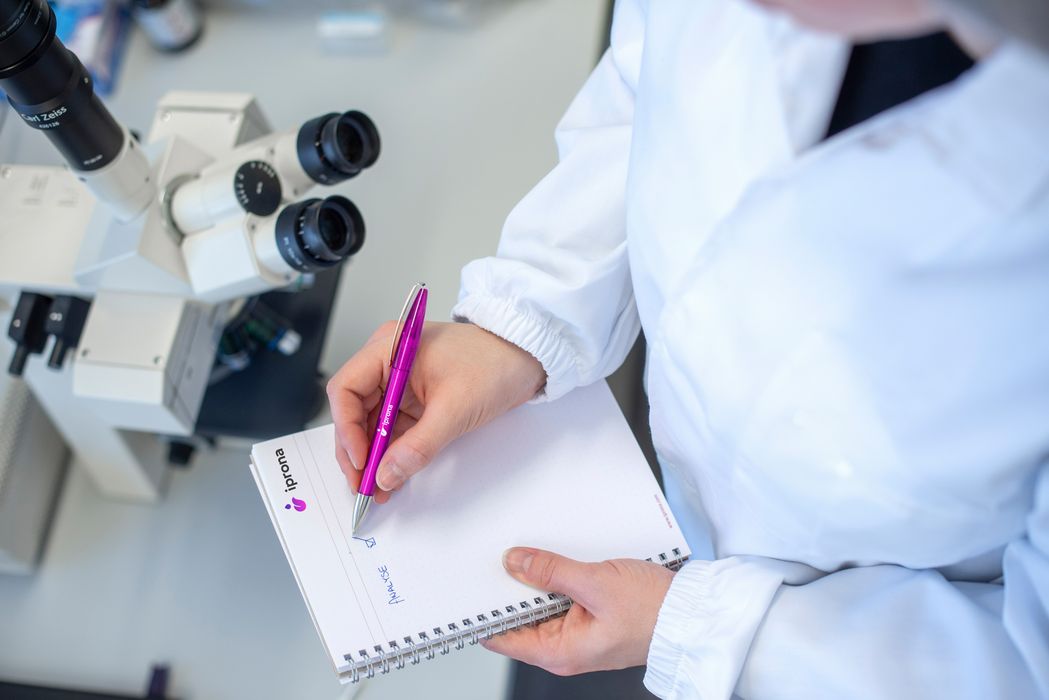 Forscher in weißem Laborkittel macht Notizen auf einem Block neben einem Mikroskop.