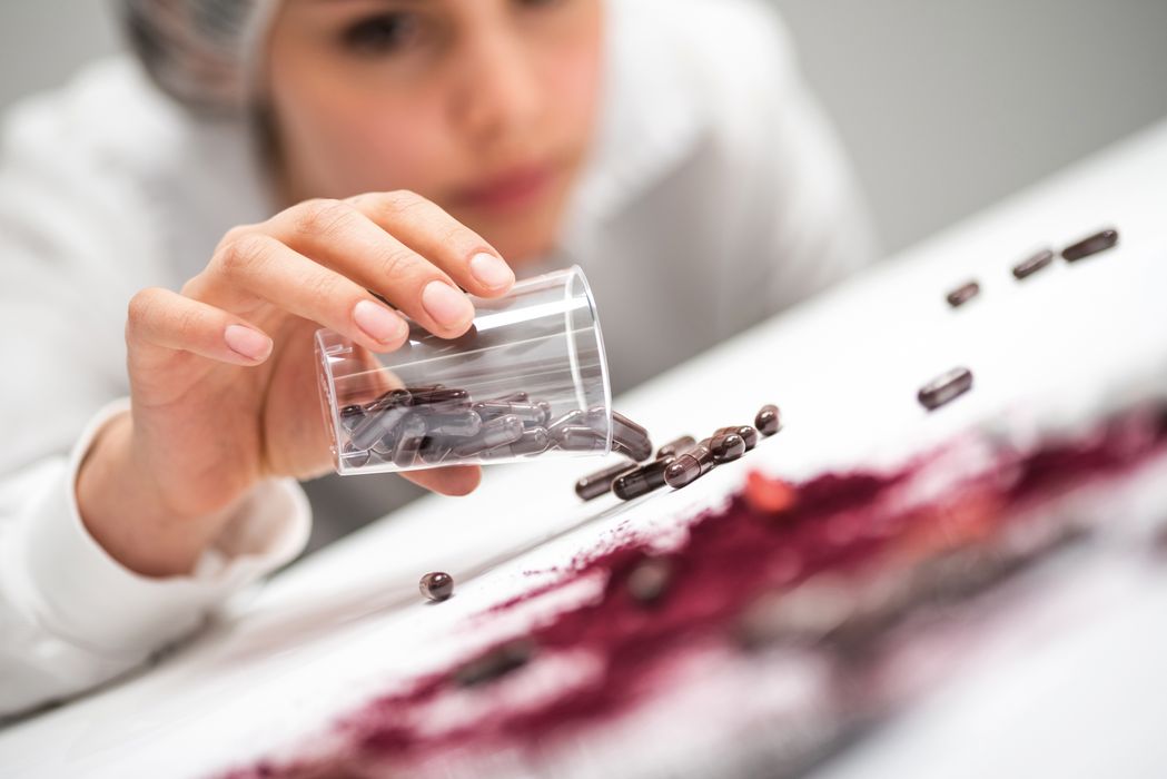 Hand gießt dunkle Kapseln aus einem durchsichtigen Becher auf eine weiße Oberfläche mit rotem Pulver.