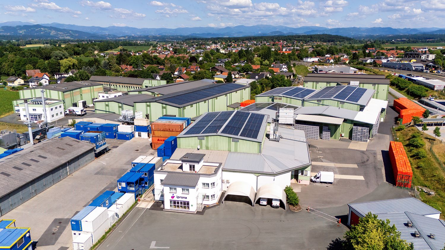 Complesso industriale con pannelli solari sui tetti, container blu e case residenziali sullo sfondo sotto un cielo parzialmente nuvoloso.