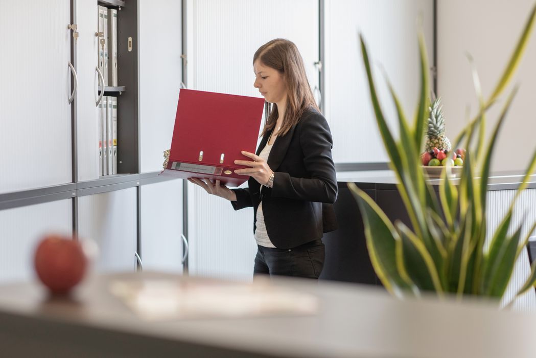Frau im Büro liest in rotem Ordner vor Aktenschränken, im Vordergrund Pflanze und Obstkorb