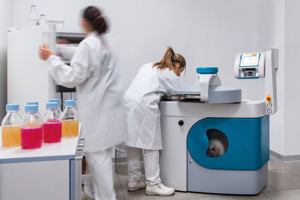 Labor mit zwei Wissenschaftlerinnen in weißen Kitteln, eine arbeitet an einem Gerät, die andere holt etwas aus einem Schrank, auf dem Tisch stehen mehrere Flaschen mit bunten Flüssigkeiten